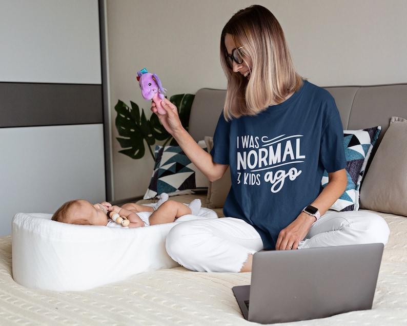 Pode incluir: Uma mulher vestindo uma camiseta azul marinho com o texto "Eu era normal h&aacute; 3 filhos" est&aacute; sentada em uma cama com um laptop. Um beb&ecirc; est&aacute; deitado em um ber&ccedil;o branco e brincando com um brinquedo roxo.