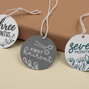 May include: Three white and gray baby milestone tags with black text and illustrations. The tags read "three months", "first haircut" with a pair of scissors, and "seven months".