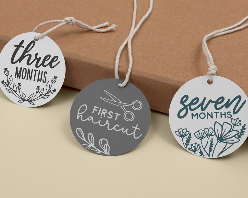 May include: Three white and gray baby milestone tags with black text and illustrations. The tags read "three months", "first haircut" with a pair of scissors, and "seven months".