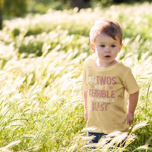 Pu&ograve; includere: Un bambino piccolo che indossa una maglietta gialla con il testo "The Twos Aren't Terrible I'm Having A Blast" in rosa. Il bambino &egrave; in piedi in un campo di erba alta.