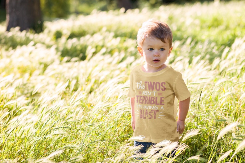 Pu&ograve; includere: Un bambino piccolo che indossa una maglietta gialla con il testo "The Twos Aren't Terrible I'm Having A Blast" in rosa. Il bambino &egrave; in piedi in un campo di erba alta.