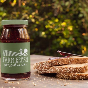 May include: A glass jar of red jam with a green label that says "FARM FRESH produce" and a white illustration of a farmhouse and windmill. The jar is sitting on a wooden table next to a stack of four slices of bread with sesame seeds.