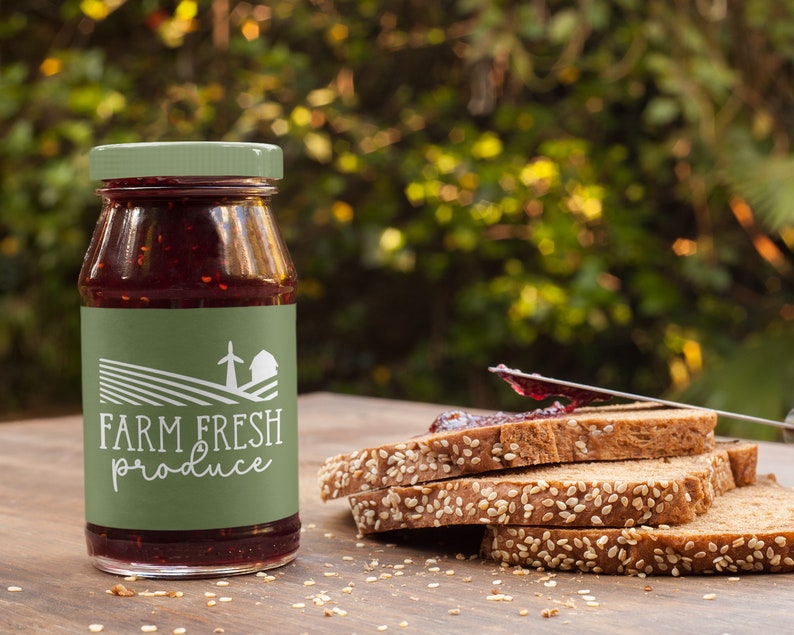 May include: A glass jar of red jam with a green label that says "FARM FRESH produce" and a white illustration of a farmhouse and windmill. The jar is sitting on a wooden table next to a stack of four slices of bread with sesame seeds.