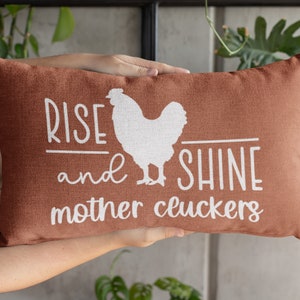 May include: A rust-colored throw pillow with white text that reads "Rise and Shine Mother Cluckers" and a white silhouette of a chicken.
