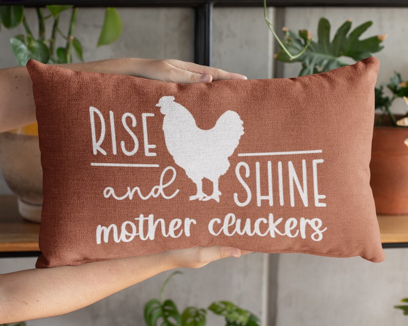 May include: A rust-colored throw pillow with white text that reads "Rise and Shine Mother Cluckers" and a white silhouette of a chicken.