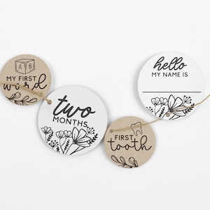 May include: A set of five round, brown and white paper milestone cards with black text and floral designs. The cards are strung together with twine and include the phrases "My First Word", "Two Months", "Hello My Name Is", and "First Tooth".