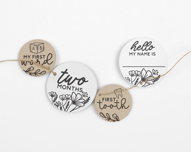 May include: A set of five round, brown and white paper milestone cards with black text and floral designs. The cards are strung together with twine and include the phrases "My First Word", "Two Months", "Hello My Name Is", and "First Tooth".