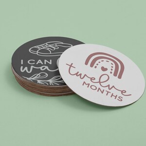 May include: A set of twelve baby milestone cards. The cards are round and feature a white background with black and pink text. The text on the cards reads "I can walk" and "twelve months".