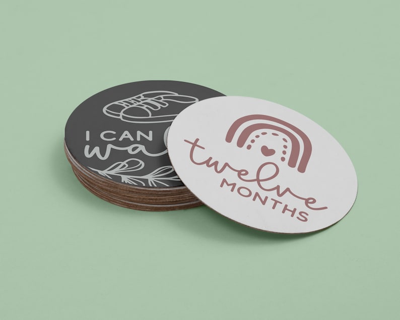 May include: A set of twelve baby milestone cards. The cards are round and feature a white background with black and pink text. The text on the cards reads "I can walk" and "twelve months".