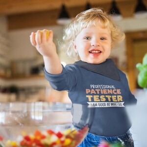 Pu&ograve; includere: Un bambino piccolo che indossa una maglietta blu a maniche lunghe con il testo "Professional Patience Tester I am good at what I do" in arancione e bianco. Il bambino sorride e tiene un limone giallo.