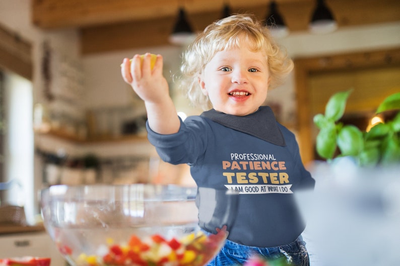 Pu&ograve; includere: Un bambino piccolo che indossa una maglietta blu a maniche lunghe con il testo "Professional Patience Tester I am good at what I do" in arancione e bianco. Il bambino sorride e tiene un limone giallo.