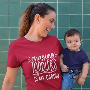 Pode incluir: Uma mulher vestindo uma camiseta vermelha com o texto "chasing toddlers is my cardio" em branco. Ela est&aacute; segurando uma crian&ccedil;a pequena que est&aacute; vestindo uma camiseta azul marinho e jeans azul.