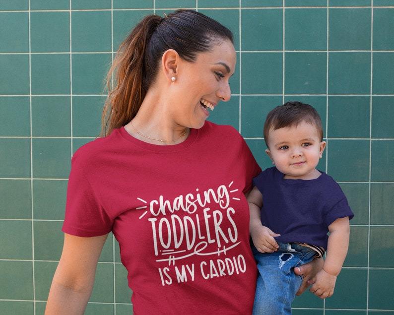 Pode incluir: Uma mulher vestindo uma camiseta vermelha com o texto "chasing toddlers is my cardio" em branco. Ela est&aacute; segurando uma crian&ccedil;a pequena que est&aacute; vestindo uma camiseta azul marinho e jeans azul.