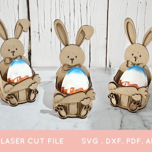 May include: Three wooden bunny rabbit Easter egg holders. Each bunny is holding a blue and orange Easter egg. The bunnies are sitting on a white surface.