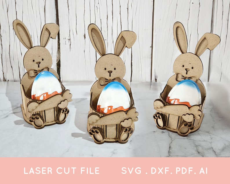 May include: Three wooden bunny rabbit Easter egg holders. Each bunny is holding a blue and orange Easter egg. The bunnies are sitting on a white surface.
