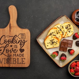 K&ouml;nnte beinhalten: Ein Holzbrett zum Schneiden mit dem Text "Cooking Love is Made Visible" eingraviert. Das Brett ist braun und hat einen Griff. Auf einem Teller liegen au&szlig;erdem verschiedene Lebensmittel, darunter Schokolade, Kuchen und Erdbeeren.