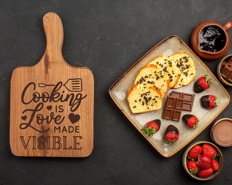 K&ouml;nnte beinhalten: Ein Holzbrett zum Schneiden mit dem Text "Cooking Love is Made Visible" eingraviert. Das Brett ist braun und hat einen Griff. Auf einem Teller liegen au&szlig;erdem verschiedene Lebensmittel, darunter Schokolade, Kuchen und Erdbeeren.