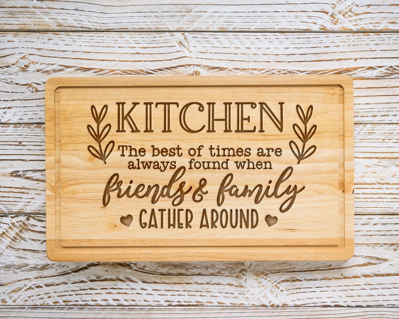 K&ouml;nnte beinhalten: Ein Schneidebrett aus Holz mit dem eingravierten Text "KITCHEN The best of times are always found when friends & family GATHER AROUND".
