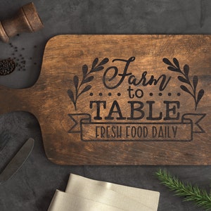 K&ouml;nnte beinhalten: Ein braunes, rechteckiges Schneidebrett aus Holz mit dem eingravierten Text "Farm to Table Fresh Food Daily". Das Brett hat einen Griff und ist mit einem Blattdesign verziert.