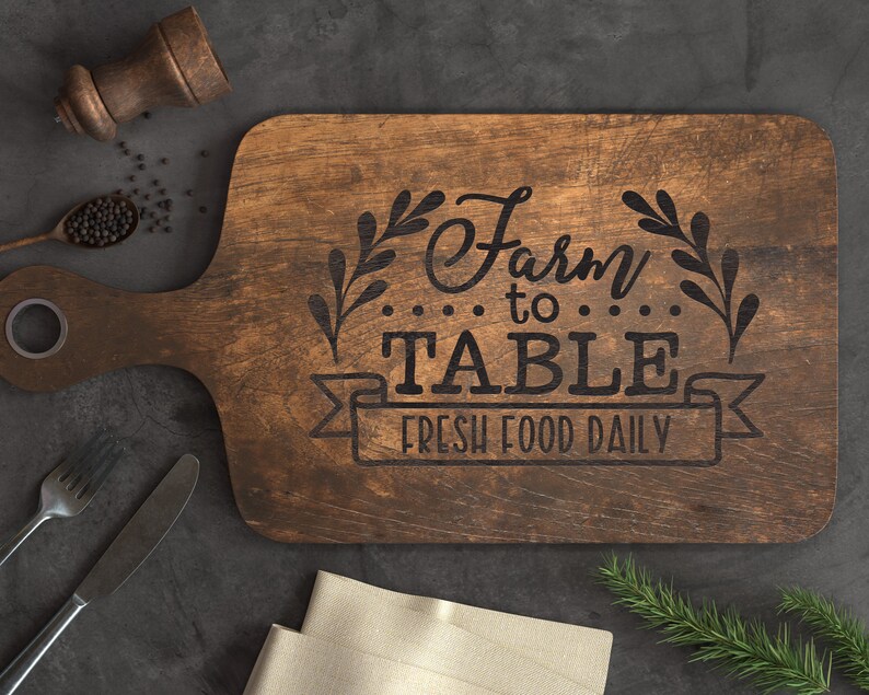 K&ouml;nnte beinhalten: Ein braunes, rechteckiges Schneidebrett aus Holz mit dem eingravierten Text "Farm to Table Fresh Food Daily". Das Brett hat einen Griff und ist mit einem Blattdesign verziert.
