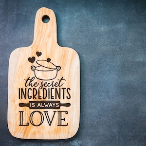 K&ouml;nnte beinhalten: Ein Holzbrett zum Schneiden mit dem Text "the secret ingredients is always LOVE" und einer Zeichnung eines Topfes mit Herzen dar&uuml;ber.