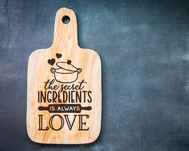 K&ouml;nnte beinhalten: Ein Holzbrett zum Schneiden mit dem Text "the secret ingredients is always LOVE" und einer Zeichnung eines Topfes mit Herzen dar&uuml;ber.
