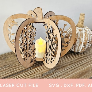 Op de afbeelding: Een lasergesneden houten pompoen-vormige lantaarn met bloemmotieven, met een batterij-aangedreven kaars. Daarnaast een decoratieve witte en bruine pompoen met een touwsteel. De tekst "LASER CUT FILE" en "SVG . DXF . PDF . AI" staat onderaan.