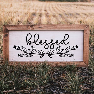May include: A wooden sign with a white background and black lettering that reads "blessed". The sign is decorated with a floral design.