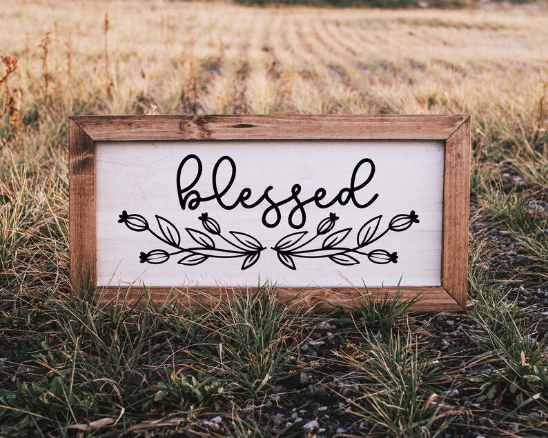 May include: A wooden sign with a white background and black lettering that reads "blessed". The sign is decorated with a floral design.