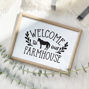 May include: A wooden framed sign with the text "WELCOME to our FARMHOUSE" in black lettering. A silhouette of a horse is included in the text. The sign is on a white wooden surface.