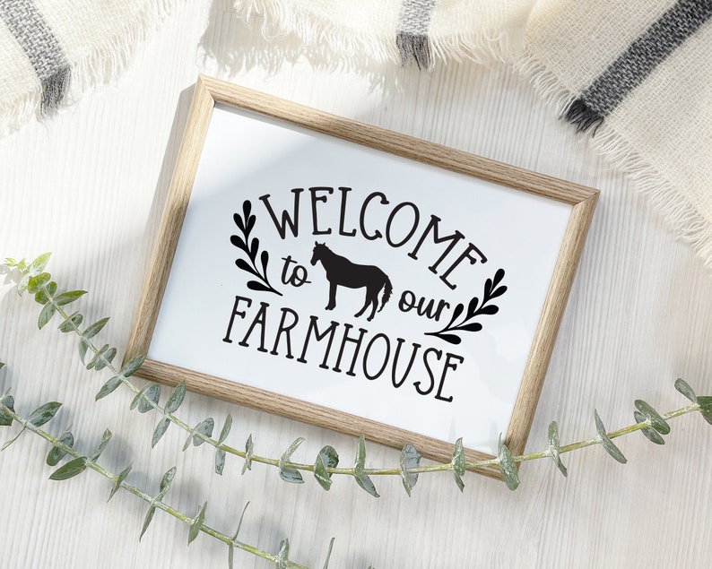 May include: A wooden framed sign with the text "WELCOME to our FARMHOUSE" in black lettering. A silhouette of a horse is included in the text. The sign is on a white wooden surface.