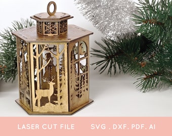 May include: A wooden laser cut lantern with a winter scene. The lantern is hexagonal and has a snowman, deer, and trees cut out of the wood. The lantern has a gold finish and is sitting on a white surface.