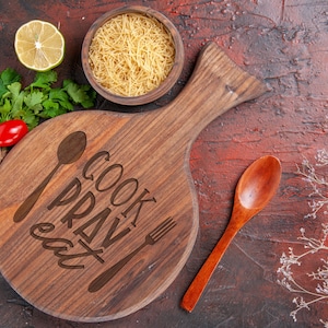 K&ouml;nnte beinhalten: Ein Holzbrett mit den Worten "Cook Pray Eat" eingraviert. Ein L&ouml;ffel und eine Gabel sind ebenfalls auf dem Brett eingraviert. Es gibt eine Sch&uuml;ssel Pasta, eine Limette und einen Holzl&ouml;ffel auf rotem Hintergrund.