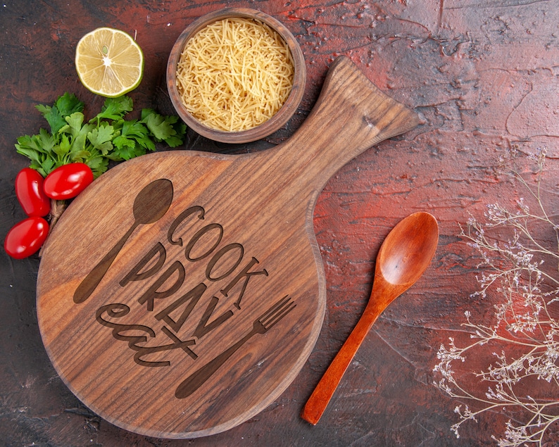 K&ouml;nnte beinhalten: Ein Holzbrett mit den Worten "Cook Pray Eat" eingraviert. Ein L&ouml;ffel und eine Gabel sind ebenfalls auf dem Brett eingraviert. Es gibt eine Sch&uuml;ssel Pasta, eine Limette und einen Holzl&ouml;ffel auf rotem Hintergrund.