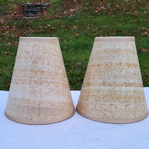 May include: Two beige lampshades with a textured, horizontal striped pattern. The lampshades have a conical shape, with a wider base and a narrower top. The top and bottom edges are trimmed with a lighter beige band.