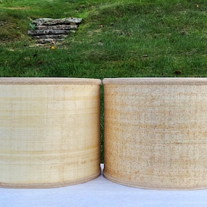 May include: Two cylindrical lampshades with a light beige woven texture. The shades have a rounded top and bottom with a darker beige trim. They are set against a backdrop of green grass and a stone wall.
