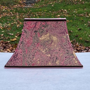 Pode incluir: Abajur retangular rosa com um padrão marmorizado em tons de rosa, dourado e preto. O abajur tem uma borda marrom e uma base rosa. O fundo mostra uma área gramada com folhas.