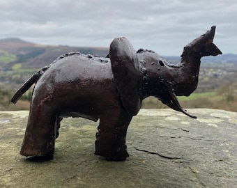 May include: A dark brown metal elephant sculpture with large ears and a raised trunk. The handcrafted elephant is standing on a stone surface, with a scenic background of rolling hills and a cloudy sky.