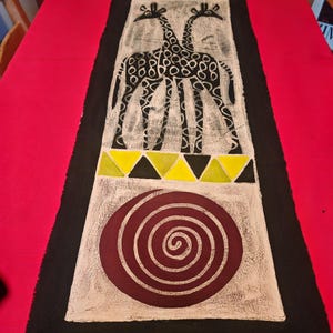 May include: A black and white table runner with a stylised design of two giraffes, a spiral, and yellow triangles. The background is a cream colour.