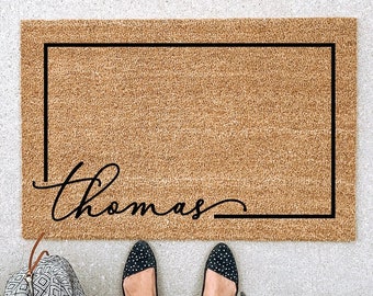May include: A rectangular doormat made of natural coir fiber, featuring a black border and the name "Thomas" in a cursive font. The mat is placed on a light gray surface, with a patterned bag and feet wearing polka dot shoes visible.