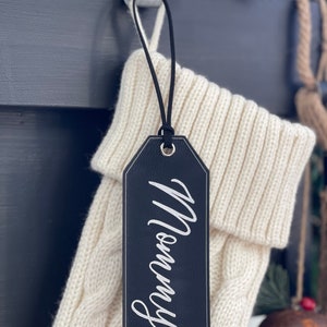 May include: A black leather stocking tag with the word "Mommy" written in white script.