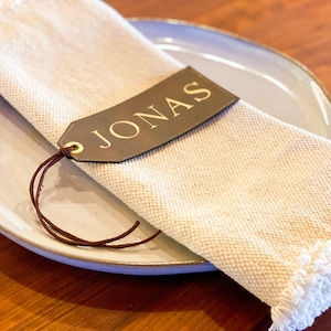 May include: A brown leather place card with the name "JONAS" written in gold lettering. The place card is tied to a white linen napkin with a brown leather string.