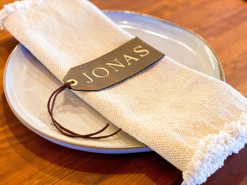 May include: A brown leather place card with the name "JONAS" written in gold lettering. The place card is tied to a white linen napkin with a brown leather string.