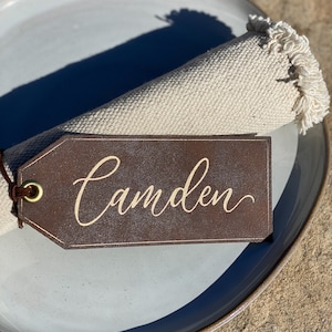 May include: A brown leather place card with the name "Camden" written in gold script. The place card is attached to a white linen napkin with a fringed edge.