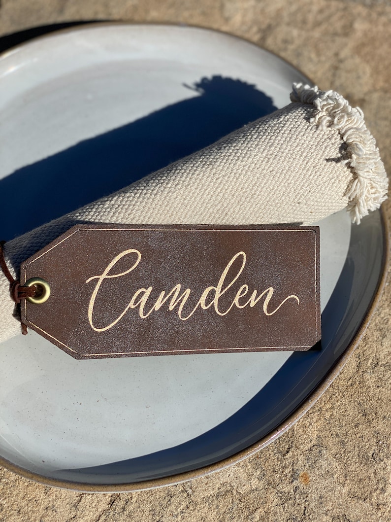 May include: A brown leather place card with the name "Camden" written in gold script. The place card is attached to a white linen napkin with a fringed edge.