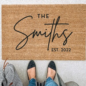 May include: A rectangular doormat made of natural fibers, featuring the text "THE Smiths EST. 2022" in black script. The mat is placed on a concrete surface, with a person's feet and a patterned bag visible.