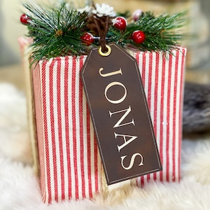 May include: A red and white striped gift box with a brown gift tag that says "JONAS" on it. The box is decorated with a sprig of pine and red berries.
