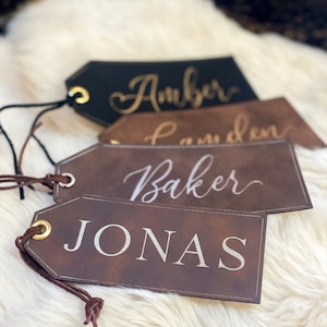 May include: Four brown leather gift tags with gold eyelets and string. The tags are labelled with the names "Amber", "Camden", "Baker", and "Jonas" in white script.