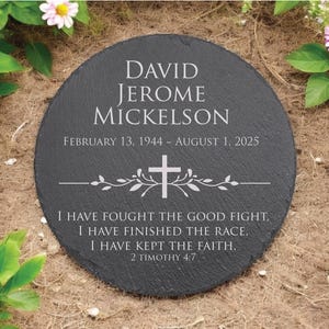 May include: A round black slate memorial stone engraved with the name David Jerome Mickelson, the dates February 13, 1944 to August 1, 2025, a central cross icon with decorative vines, and the scripture text I have fought the good fight.