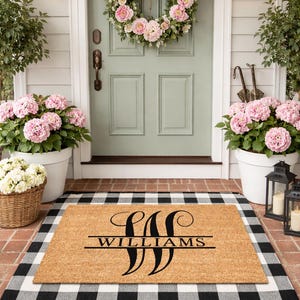 May include: A personalized doormat with the name "WILLIAMS" in black script, framed by a black and white checkered border. The mat sits on a brick surface, with potted pink flowers and a floral wreath on the door.